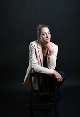 Naklejka premium Stimmungsvolles Frauenportrait im Low-Key-Licht. Selbstbewusstes Model mit Blazer auf Stuhl vor dunklem Hintergrund – modernes Business- und Charakterportrait mit Tiefe.