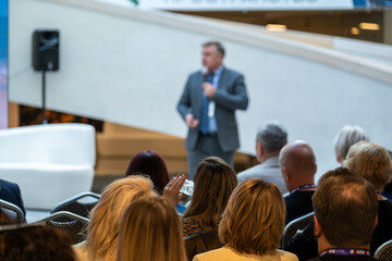 Fototapeta premium Speaker in suit delivers talk to attentive audience in modern venue. Audience listens closely with notebooks and smiles, creating professional atmosphere.