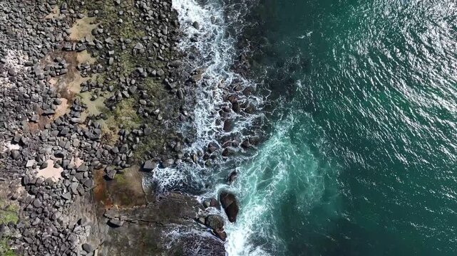 Blue water crashes against rugged rocks on a sunny day, white foam spraying high as bright blue skies reflect the golden sun, shimmering across the sparkling sea and dancing in the radient light.
