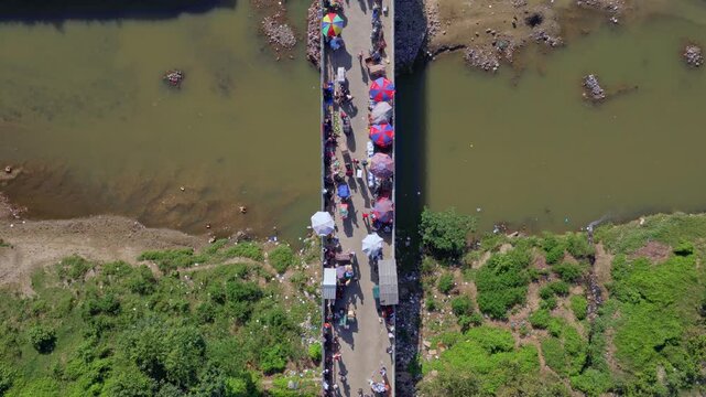 Top-down aerial view of Dominican&ndash;Haitian border in Dajab&oacute;n. Crowds gather along narrow bridge over river as vendors and traders prepare for busy cross-border market day under bright sun.