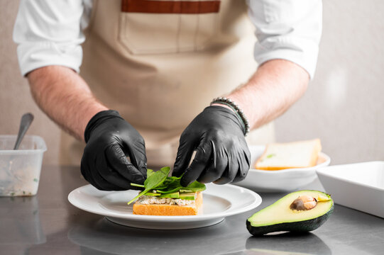 A professional chef wearing black nitrile gloves carefully garnishes a healthy avocado and tuna open-faced sandwich with fresh baby spinach leaves in a bright, modern commercial kitchen setting