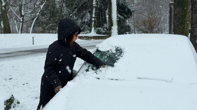 Woman in black winter jacket removing thick snow from parked car windshield during active snowfall. Real winter driving preparation scene in snowy Slovakia, cold weather lifestyle concept.