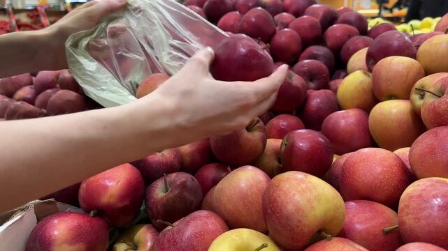 Woman choosing fresh apples in grocery store, customer picking fruits into plastic bag in supermarket produce section, healthy food shopping concept 4K