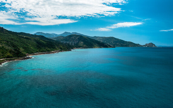 Aerial view of Sicily's north coast near Finale, with turquoise sea, rugged rocky cliffs, a coastal viaduct and distant Rocca under a cloud-dotted sky. Finale, Sicilia, Italy.