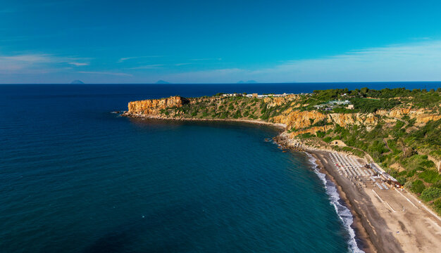 Aerial view of Capo Raisigerbi on Sicily's north coast, with Spiaggia Torre Conca, rugged cliffs, dark sand and turquoise Mediterranean water in a wide panoramic landscape