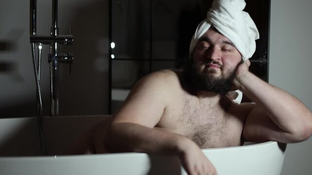 Defocused horizontal 4K video. Naked sad fat white man with towel on head sitting in bathtub in dark hotel bath room. 8 march, purity, resort, men day, rest, spa, relax, mental health, wash, skin care