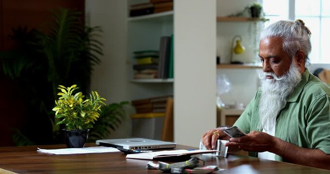 Indian mid age man counting Indian money notes at home, stylish bearded male managing monthly finance using calculator and diary while handling five hundred rupee currency notes at table