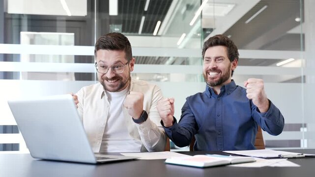 Two happy excited coworkers watching online results or cheering for bids at auction using laptop sitting in business office. Joyful businessmen celebrating success showing excitement and achievement