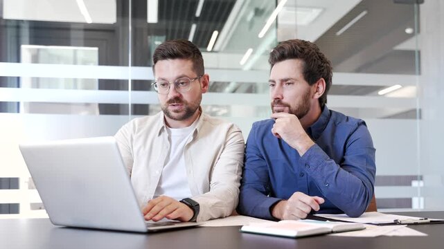 Happy businessman shows colleague great results on laptop computer while sitting at desk at workplace in a modern business office. Joyful worker brags to friend about successful work and achievements