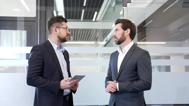 Two confident businessmen talking communicating while standing in modern office. Colleagues in suits engaged in teamwork, discuss work issues. Professionals exchanging ideas for project development