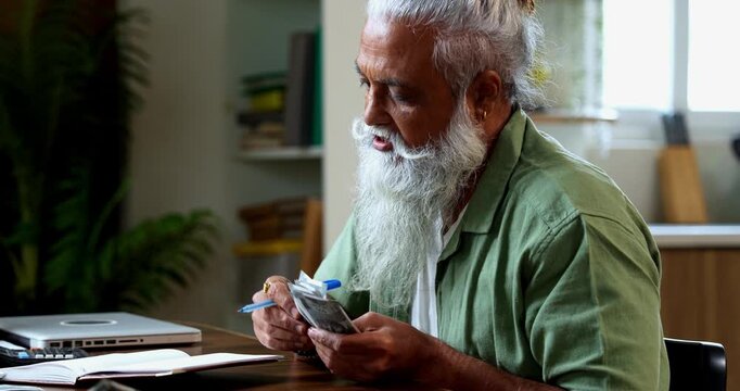 Indian mid age man counting Indian money notes at home, stylish bearded male managing monthly finance using calculator and diary while handling five hundred rupee currency notes at table
