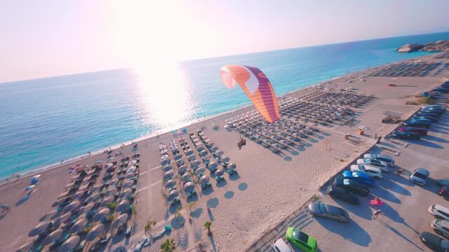 Cinematic FPV drone flight approaching a paraglider soaring above the coastline of Lefkada, Greece. Bright summer conditions reveal turquoise sea, sandy beach, and dramatic cliffs as the pilot glides 