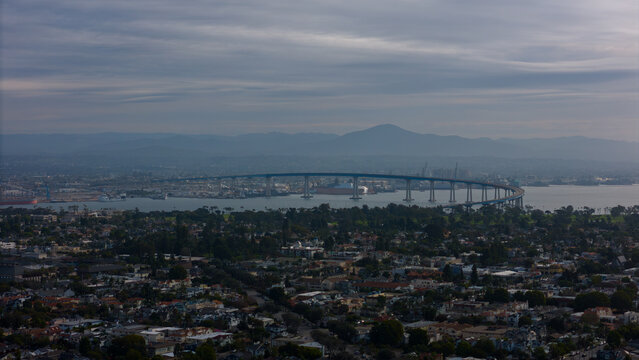 Aerial view of the Coronado Bridge arcing gracefully over the bay, connecting the city skyline with distant peaks under a soft, diffused light., Coronado, California, United States.