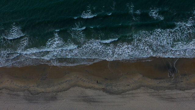 Aerial view of the dark, turbulent ocean waves crashing onto the sandy beach, creating a mesmerizing contrast of colors and textures, Coronado, California, United States.