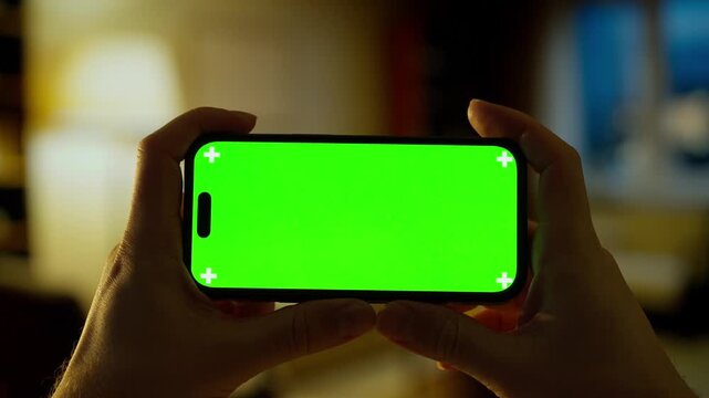 Hands holding modern smartphone with vibrant green screen in cozy dimly lit room, perfect for compositing, mockups, tutorials, or digital design presentations