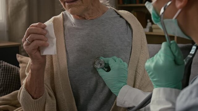 Midsection shot of coughing elderly woman holding piece of tissue visited by home care specialist wearing uniform and rubber gloves, and getting her lungs checked with stethoscope
