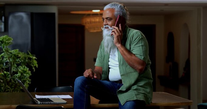 Indian mid age man speaking on phone and using laptop while sitting on table top at home, stylish bearded professional engaged in business talk through smartphone in modern interior