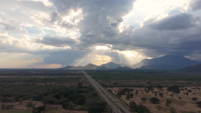 Aerial descending shot moving from a high vista to tree-level, following a gray pickup truck on a straight Colombian highway. Features dramatic sunbeams over the Cesar valley mountains at dusk.