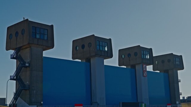 A detailed perspective shot focusing on the upper concrete control rooms and circular windows of the Blue Iwabuchi Sluice Gate. The massive blue floodgate panel is seen below against a clear, soft sky