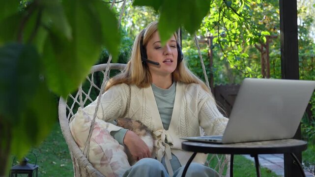 Middle aged woman working remotely with laptop and cat on her lap on terrace. Warm light, cozy lifestyle, family emotions, peaceful outdoor home office vibe.