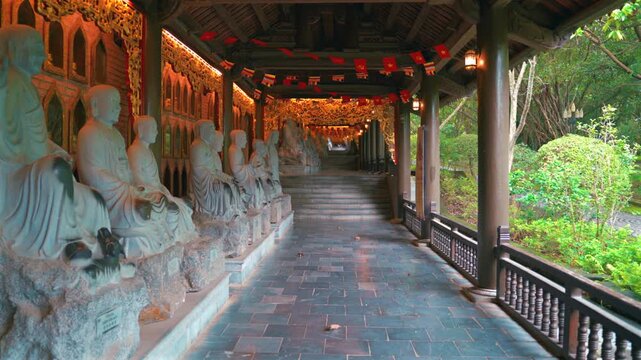 A long, ornate wooden corridor lined with numerous white stone Arhat statues leading up a staircase, decorated with Buddhist flags and warm lanterns at the Bai Dinh Pagoda in Ninh Binh, Vietnam.