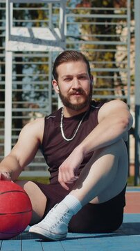 Portrait of tired athletic Lebanese man in sportswear after playing basketball game. Boy sitting relaxing on urban city court, listening music, holding ball. Fitness routine on sports field. Outdoors