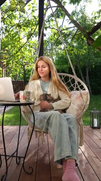 Middle aged woman working remotely with laptop and cat on her lap on terrace. Warm light, cozy lifestyle, family emotions, peaceful outdoor home office vibe.