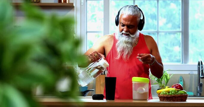 Indian mid age fit man with beard making protein shake post workout at home, stylish healthy male wearing headphones and sando vest preparing nutrition supplement in modern kitchen interior