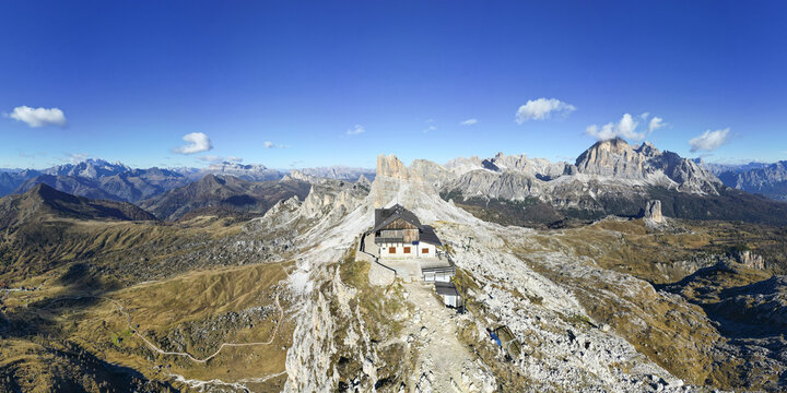 Aerial view of Rifugio Averau perched atop rugged peaks amidst the Dolomites' grandeur, a stark contrast against the azure sky, Passo Giau, Veneto, Italy.