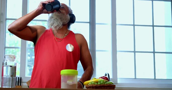 Indian mid age fit man with beard making protein shake post workout at home, stylish healthy male wearing headphones and sando vest preparing nutrition supplement in modern kitchen interior