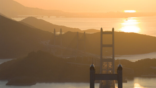 Aerial view of the Tsing Ma Bridge piercing through the golden haze as the sun casts a warm glow over the landscape, Hong Kong, Hong Kong.