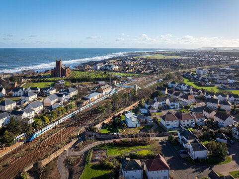 Aerial view of a train glides past the coastal town, with the North Sea stretching to the horizon and the historic church standing proudly, Dunbar, Scotland, United Kingdom.