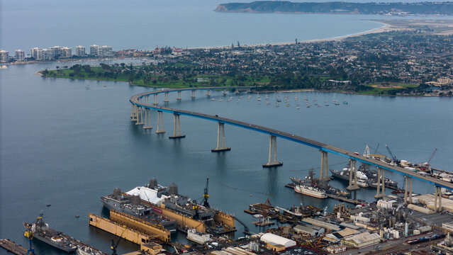 Aerial view of the Coronado Bridge stretching across sparkling waters towards the city skyline, with naval ships docked below, San Diego, California, United States.