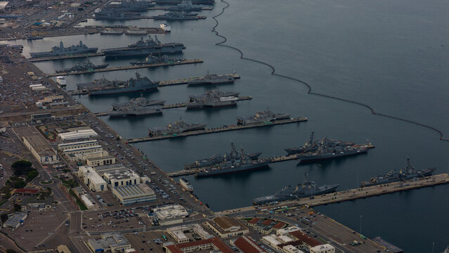 Aerial view of a fleet of naval ships docked along the harbor, contrasting with the adjacent city buildings and streets, San Diego, California, United States.