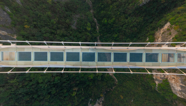 Aerial view of the Glass Bridge cutting a stark, geometric line through the rugged green mountains below, a dance of modern engineering and untouched nature, Glassbridge, Zhangjiajie, China.