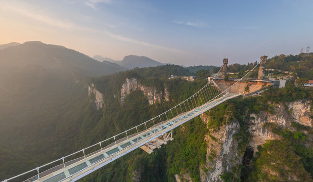 Aerial view of a long, transparent glass bridge stretching across a lush, green valley, contrasting with the rough, rocky cliffs, Glassbridge, Zhangjiajie, China.