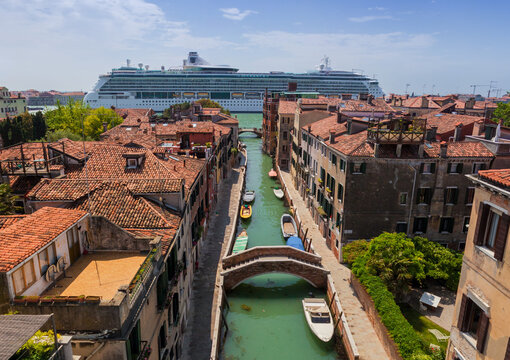Aerial view of terracotta rooftops cascading down to narrow canals, where gondolas and boats glide beneath ancient stone bridges, with a cruise ship in the background, Venice, Veneto, Italy.