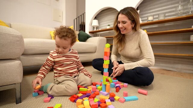 Mother and toddler son building a colorful tower with wooden blocks at home