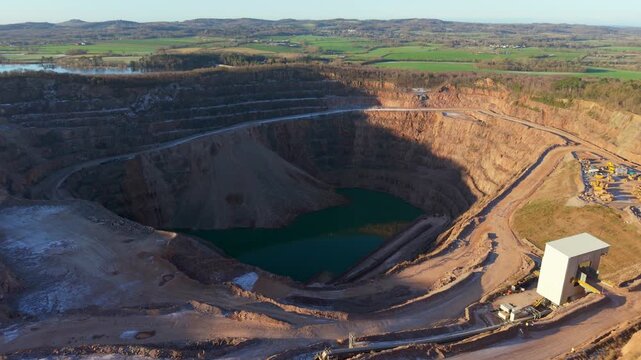 Aerial quarry excavation revealing deep open pit filling with green water. Expansive mining landscape exposing terraced rock walls descending toward flooded bottom. Massive open pit mining area