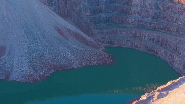 Steep quarry basin filling with deep green water beneath layered rock walls. Curved open pit excavation collecting water along terraced mineral slopes. Sunken mining cavity forming artificial lake