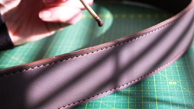 Close-up of a craftsman carefully applying edge paint to a leather belt. Precise handwork in warm natural light highlights traditional leather craftsmanship and attention to detail.