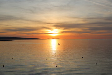 Obraz premium Beautiful golden sunset over a calm sea with a small boat silhouette on the horizon. Dramatic sky with warm orange and yellow tones reflecting on smooth water surface