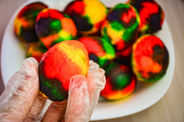 Gloved hands hold a brightly dyed Easter egg with rainbow colors. Blurred plate of painted eggs enhances festive spring mood. Perfect for holiday ads, greeting cards, blogs, and seasonal promotions.