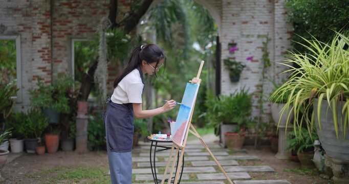 asian woman artist painting on canvas outdoors in garden while standing near stone path wearing casual clothes and apron expressing peaceful creative lifestyle in quiet backyard