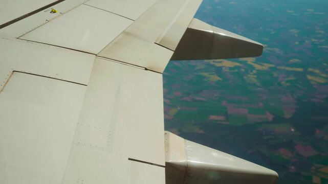 Plane flying over farmland, showing flaps, slats, and spoiler. Aircraft cruising above fields. Airplane soaring high with visible slats, flaps, and spoiler