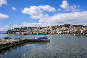 Vibrant panorama of Ohrid's historic lakeside district under a bright blue sky with fluffy clouds, featuring traditional houses, an ancient pier, and a charming overturned blue boat.
