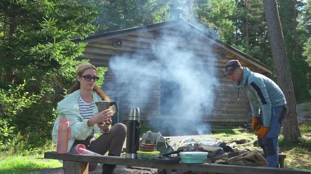 Young couple using designated campfire site in Finnish national park. Safe outdoor cooking, sustainable tourism infrastructure and responsible nature recreation concept in sunny Nordic forest.