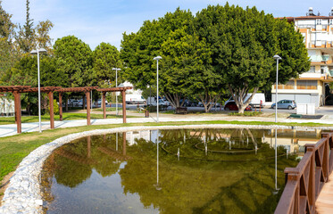 Obraz premium Modern city park with a decorative pond, wooden pergolas and lush green trees. Urban landscaping and public recreation area in Manavgat, Antalya province, Turkey. Sunny autumn day scenery.