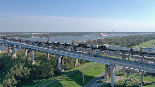 Aerial view of a freight train traversing a bridge over a wide river, connecting industrial areas under a clear sky, New Orleans, USA.