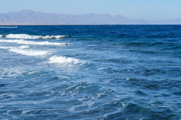 Obraz premium Ocean waves gently rolling onto a sandy beach with distant mountains visible under a clear blue sky in a coastal landscape